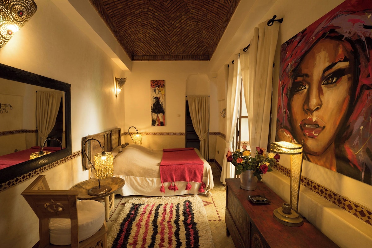 A hotel room with a bed, a mirror, a painting, a lamp, and a vase with flowers.
