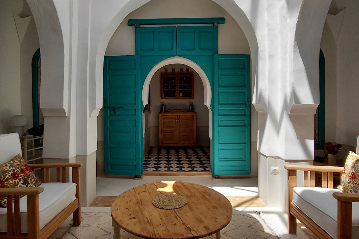 An interior space with a wooden table, two sofas, a kitchen area with a tiled floor, and a teal-colored door.