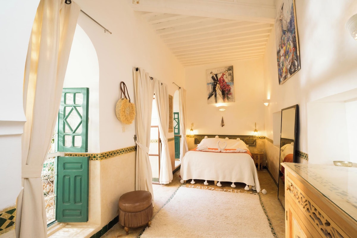 A hotel room with a bed, a small stool, a mirror, and a painting on the wall.