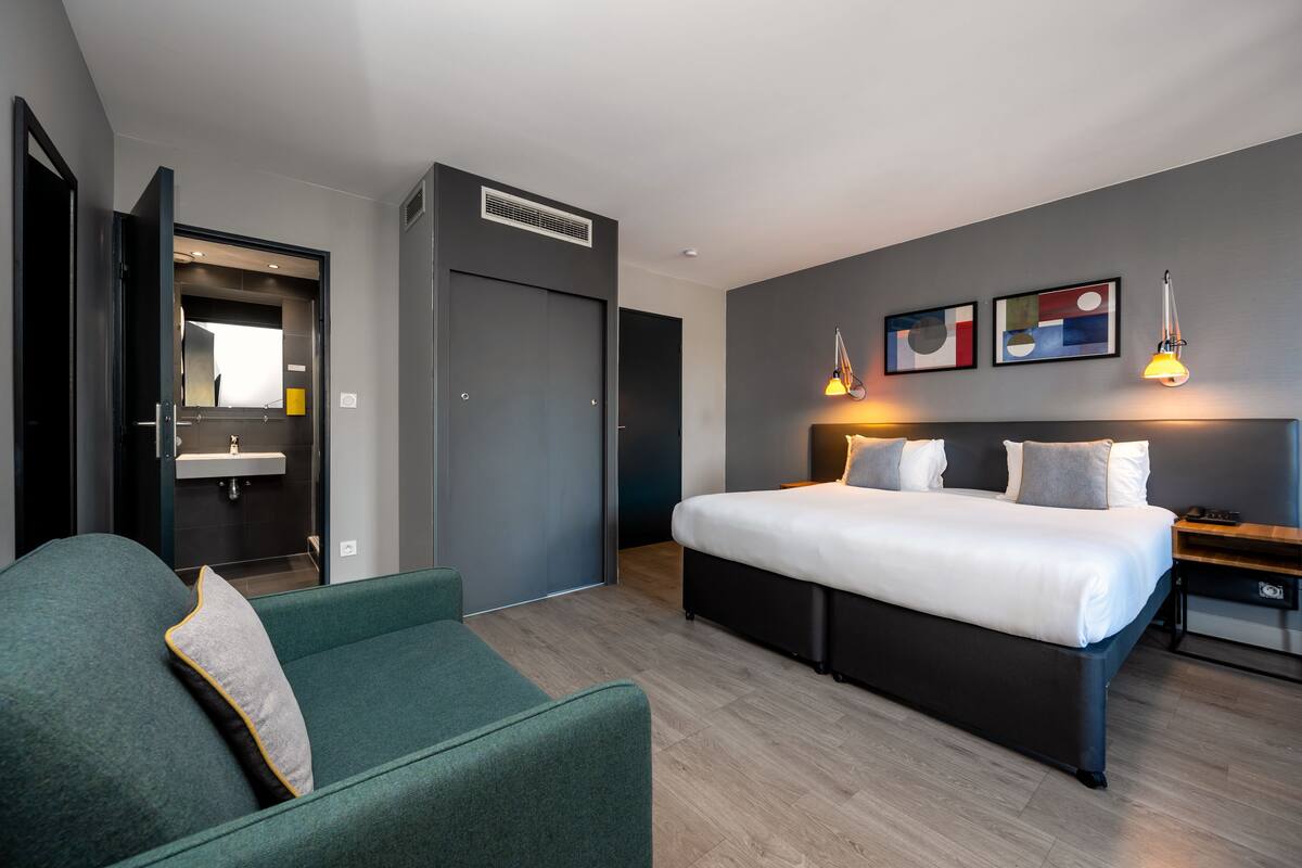 Une chambre d’hôtel moderne avec un grand lit, un canapé vert, une table de chevet et une lampe fixée au mur.