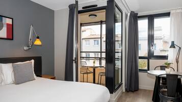 Quarto de hotel com uma cama, uma escrivaninha com cadeira, varanda com mesa e cadeiras e vista para edifícios.