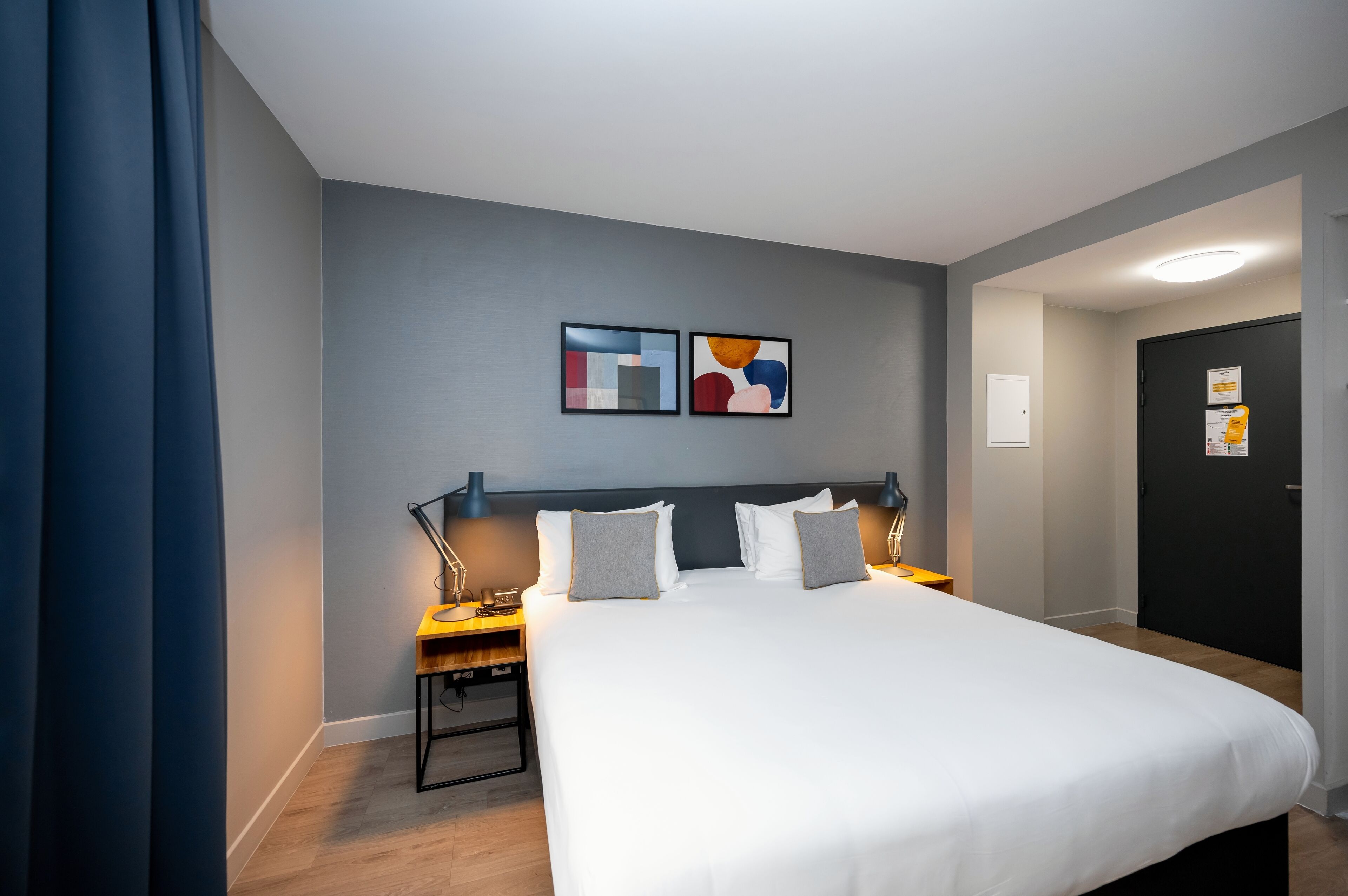 Een hotelkamer met een groot bed, twee nachtkastjes met lampen, een donkere deur en twee abstracte schilderijen aan de muur.