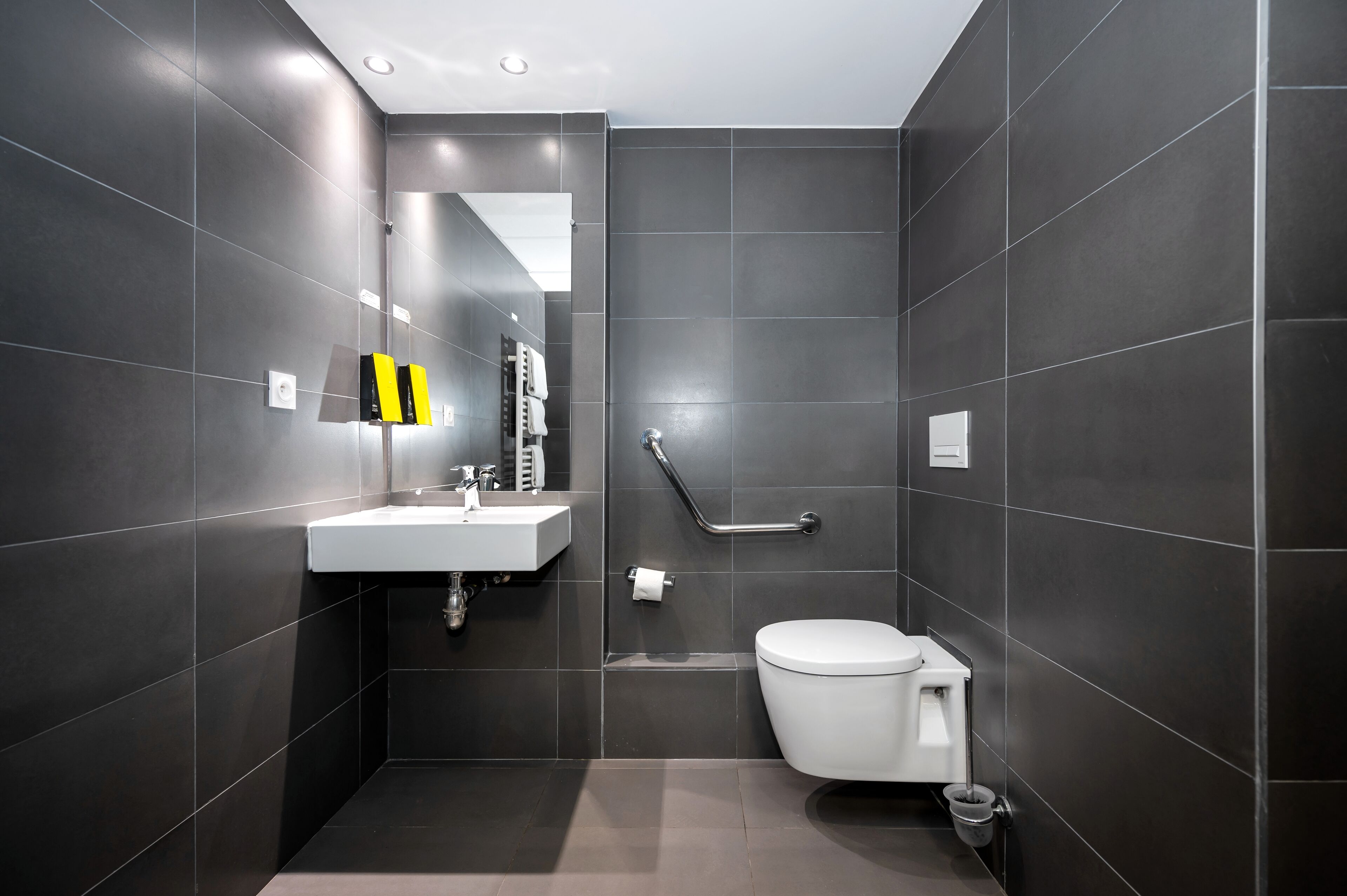 Een moderne badkamer met donkere tegels, een wit wastafelmeubel, toilet en een handgreep.