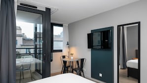 Une chambre d’hôtel moderne comprenant un lit, une petite table ronde avec deux chaises, un téléviseur fixé au mur et un balcon avec une chaise et une table.