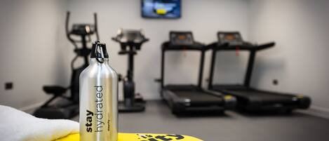 A gym with treadmills and exercise equipment, a water bottle on a yellow towel, and a TV on the wall.