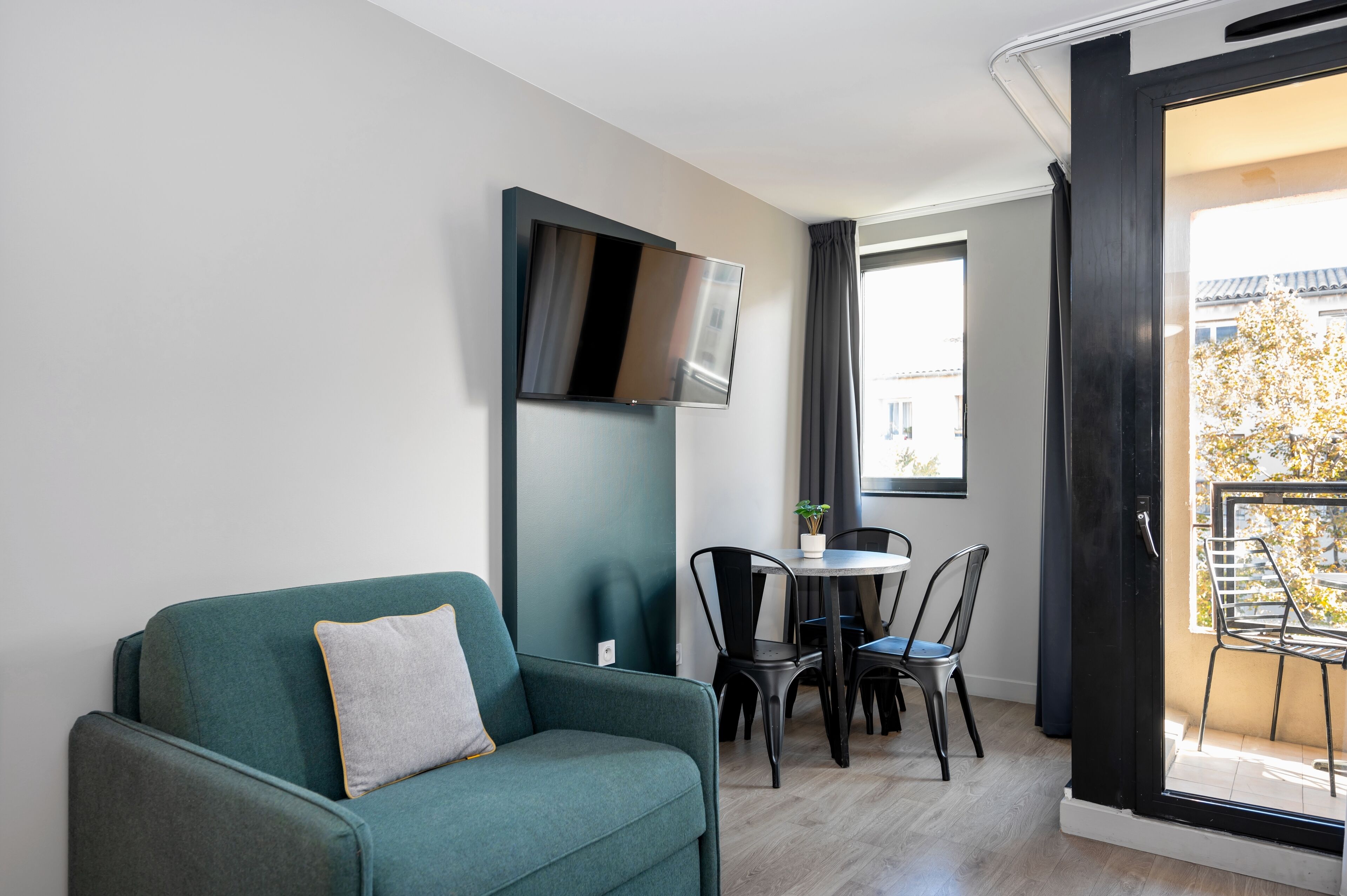 Una sala moderna con un sofá color turquesa, un televisor de pantalla plana montado en la pared, una pequeña mesa redonda con sillas, y un balcón con vistas a edificios y árboles.