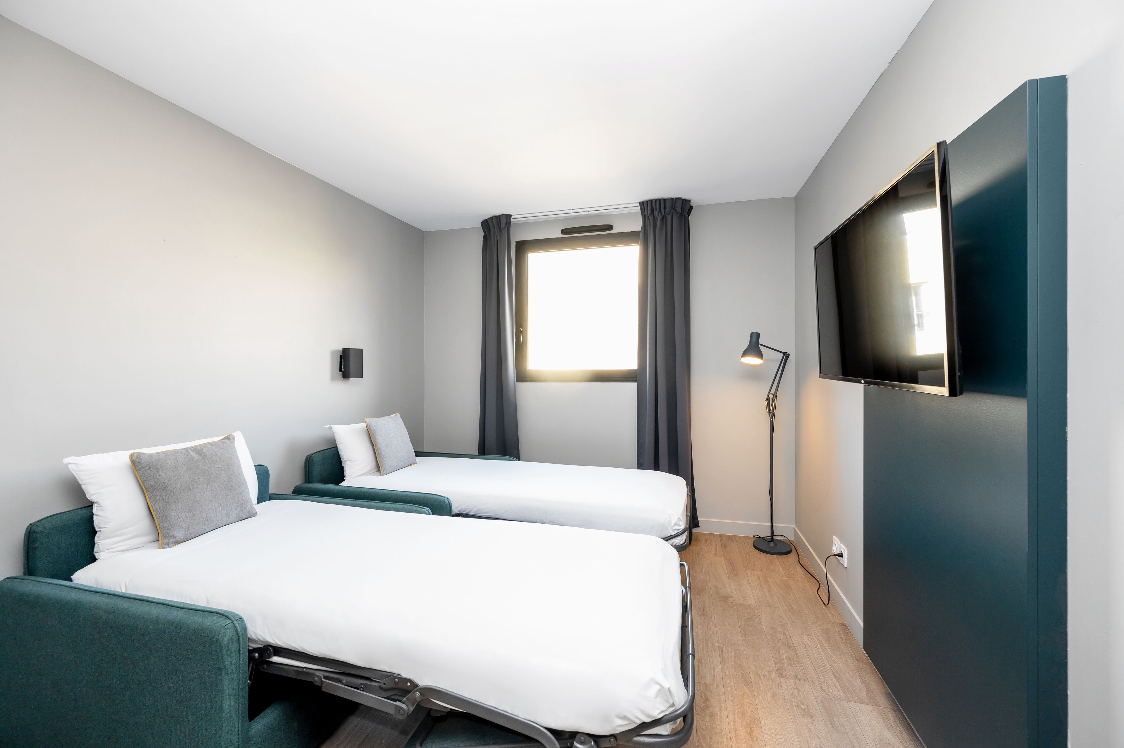 A hotel room with two beds, a TV, a window with curtains, and a floor lamp.