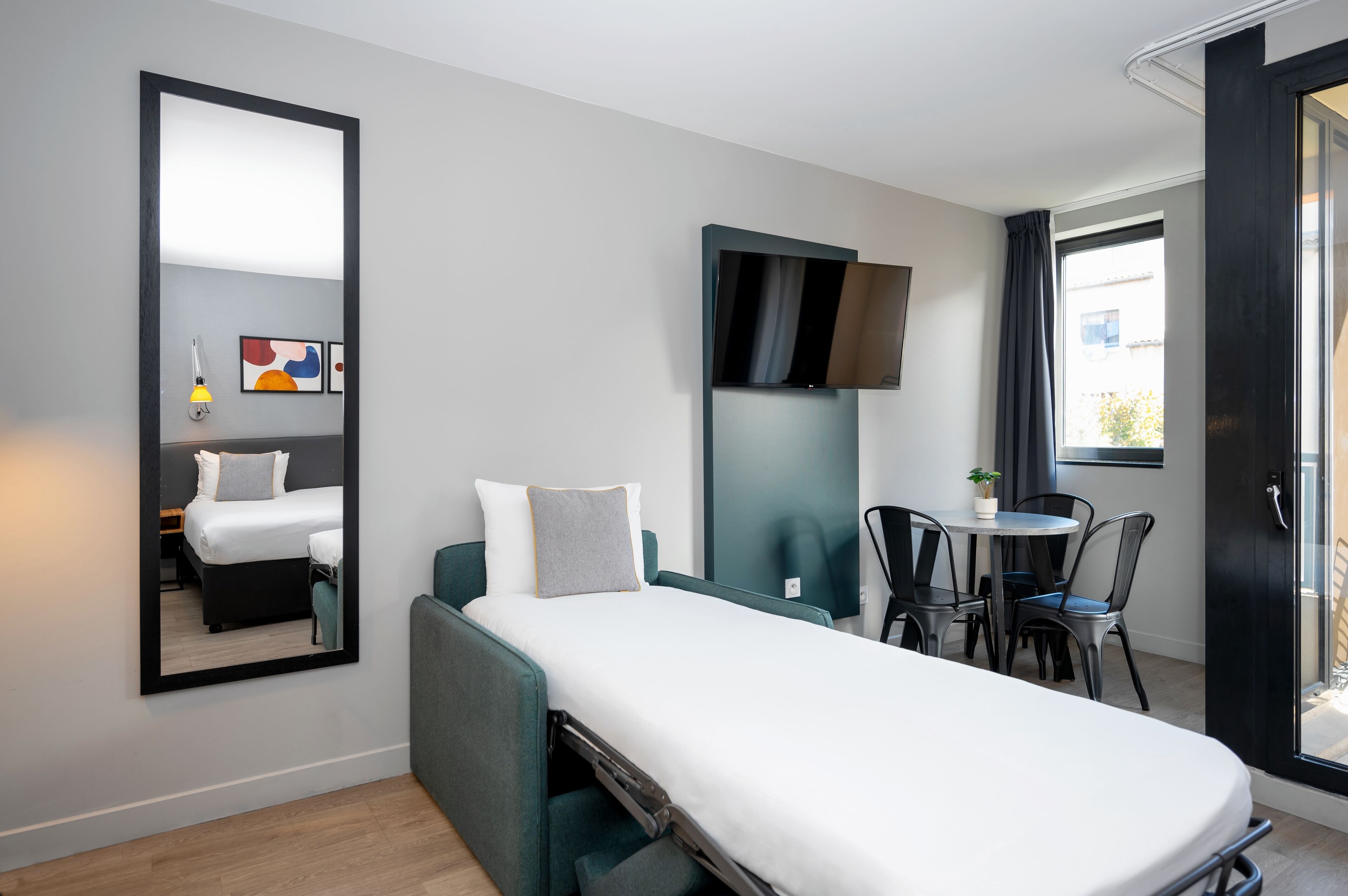 Habitación de hotel pequeña con una cama individual, una mesita con sillas, un televisor montado en la pared y un espejo grande.