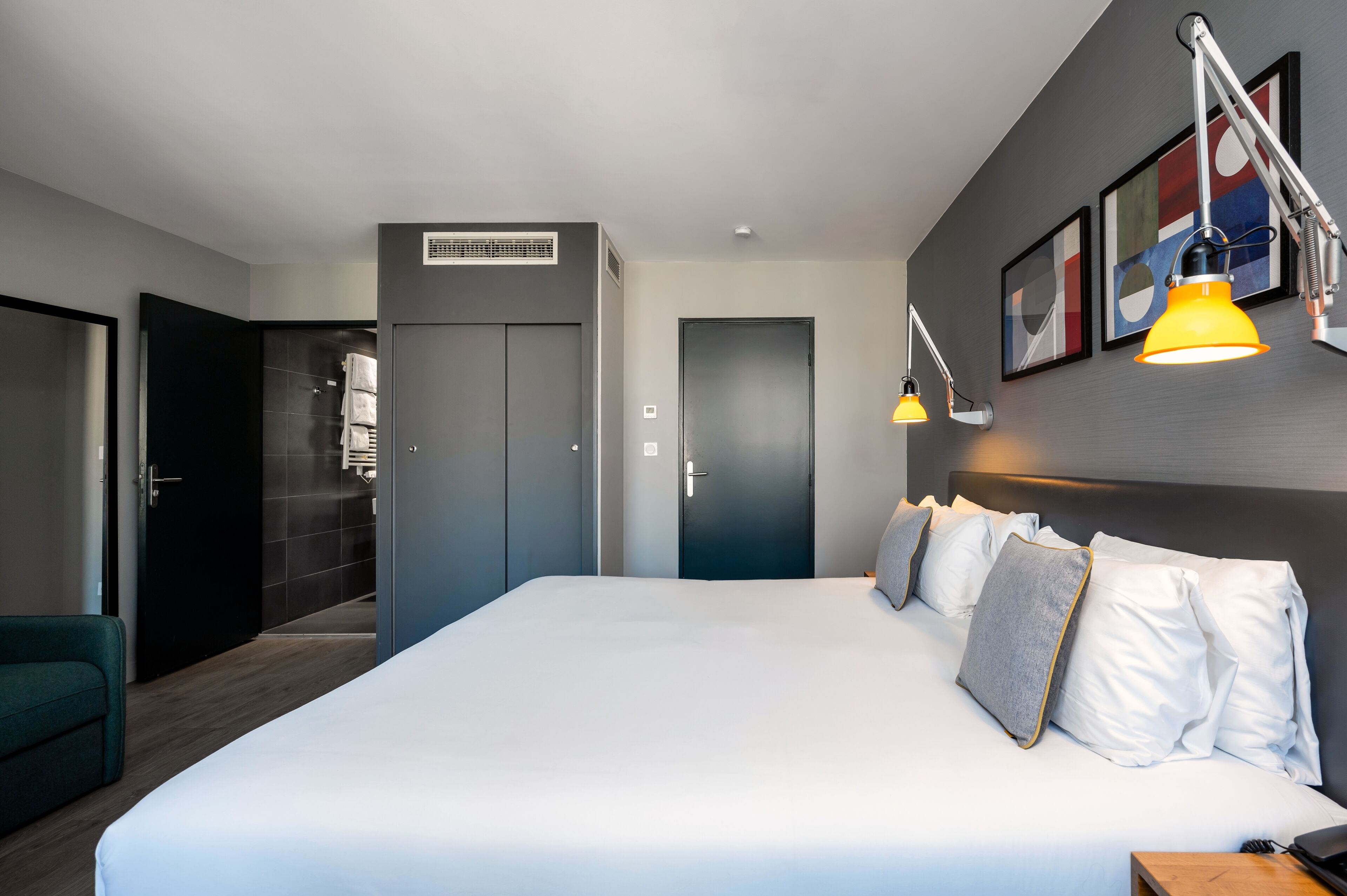 A modern hotel room with a large bed, a desk, and a chair. There are two doors, one leading to a bathroom and another to a closet.