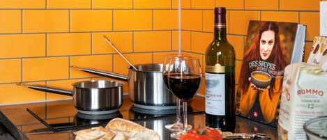 A kitchen counter with cooking pots, a wine bottle, a glass of red wine, bread, tomatoes, a cucumber, and a book titled "DES SOUPS".