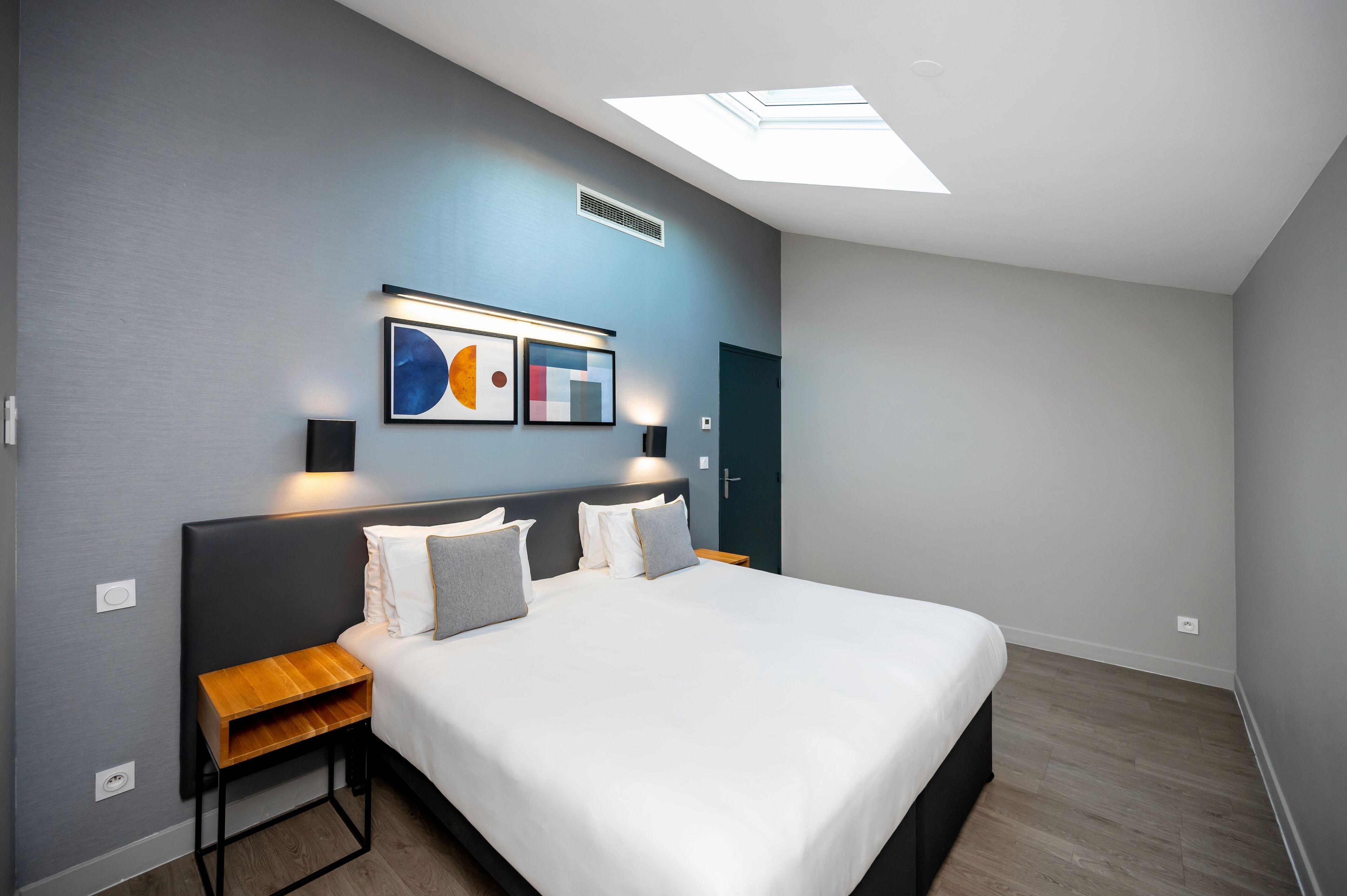 A modern bedroom with a skylight, a bed with white linens, a wooden nightstand, and abstract wall art.
