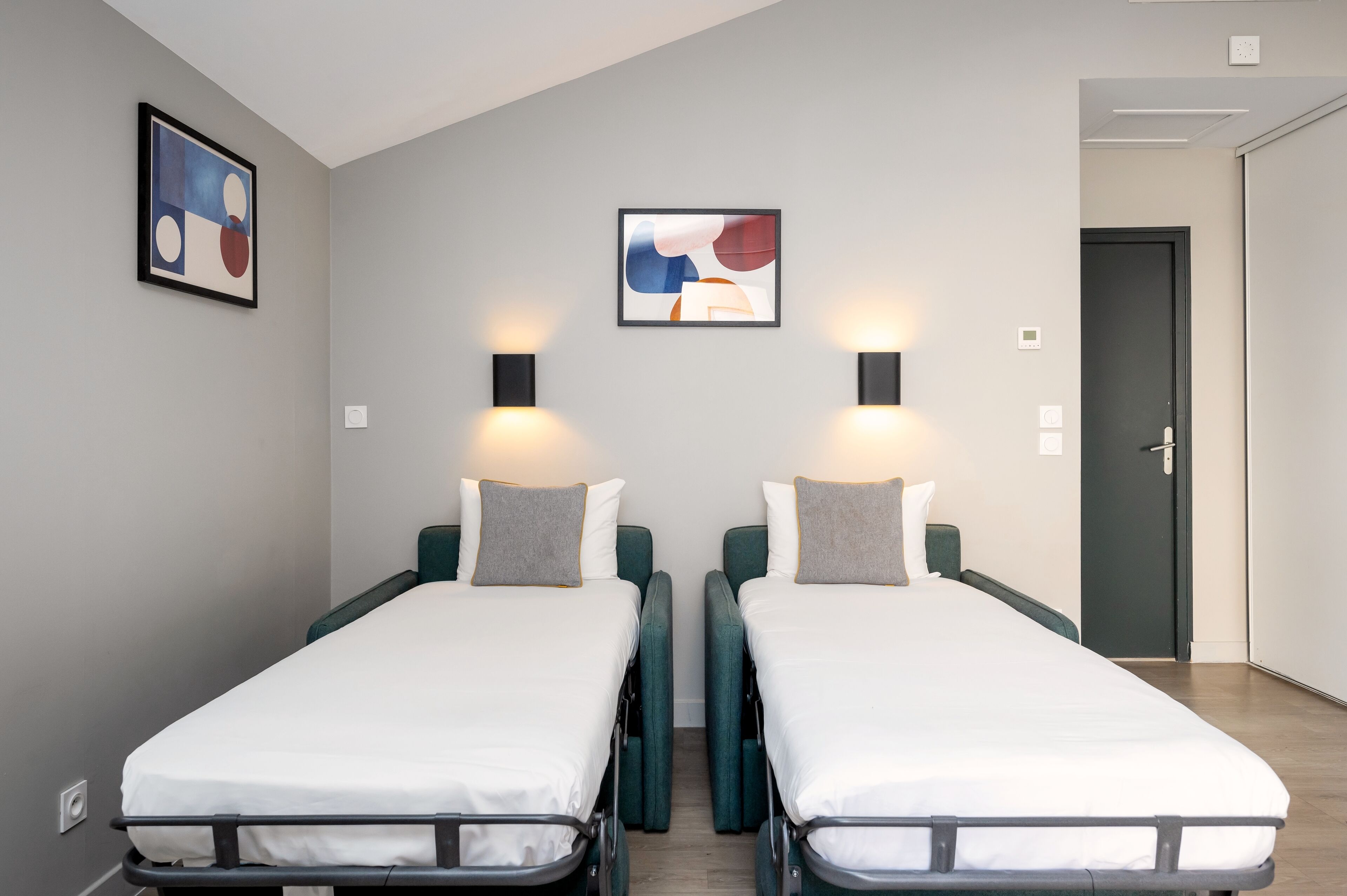 A room with two single beds, each with a grey pillow and a white mattress. There are two wall-mounted lamps on either side of the beds.