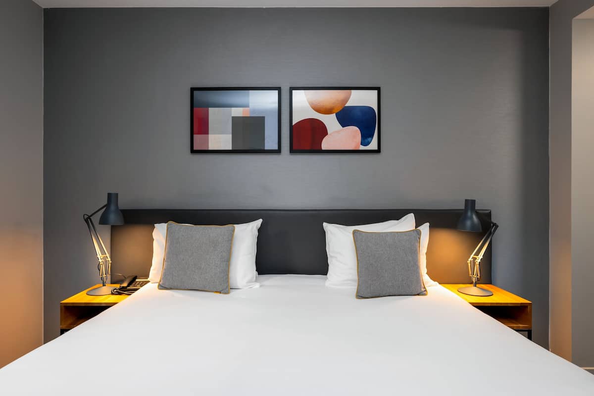 Une chambre d’hôtel moderne avec un grand lit, deux tables de chevet avec des lampes et une œuvre d’art abstraite au mur.