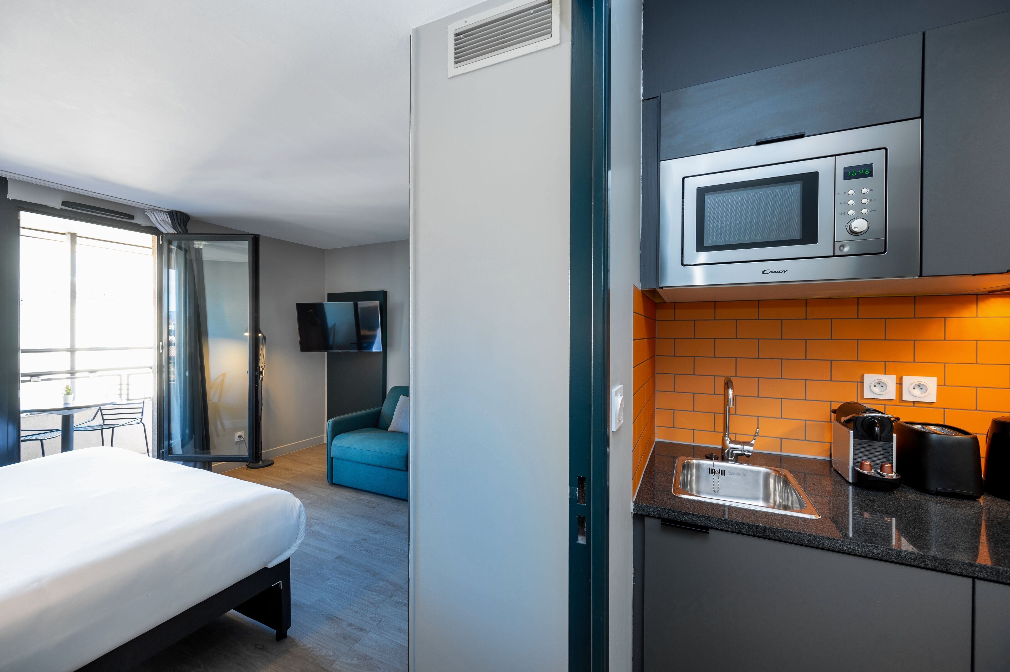 Ein kompaktes Hotelzimmer mit Bett, einer Küchenzeile inklusive Mikrowelle, Spüle und einer kleinen Essecke mit Tisch und Stühlen.