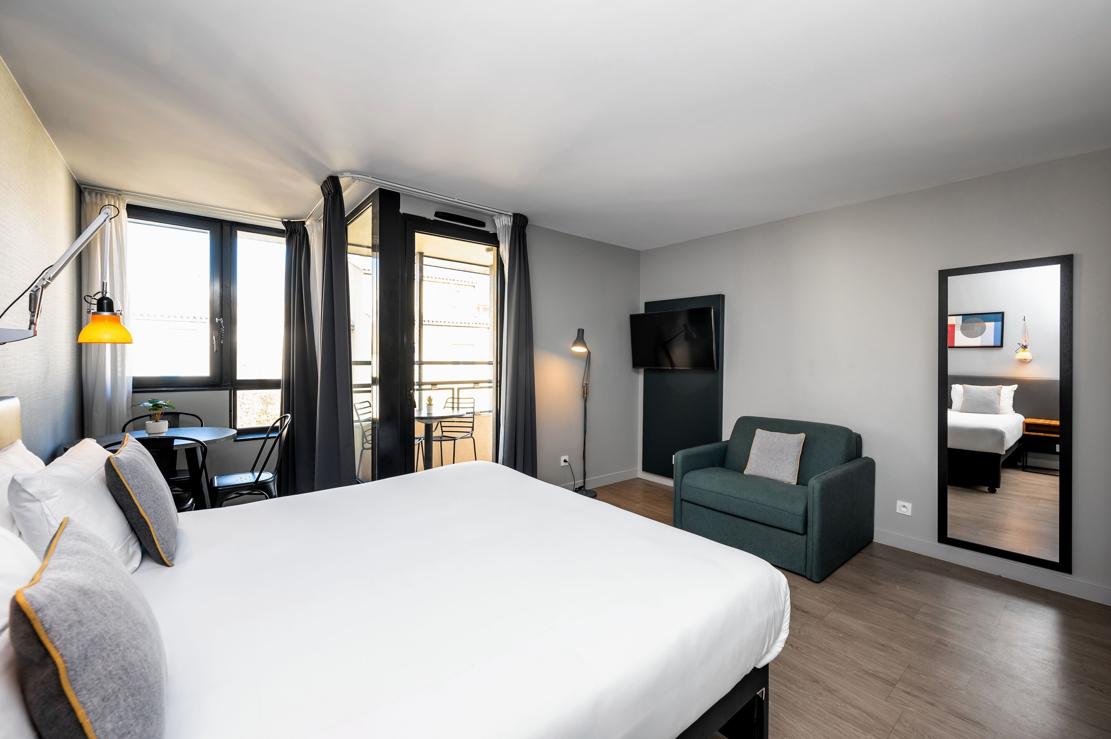 A modern hotel room with a large bed, a desk with a chair, a sofa, a TV, and a balcony with curtains.
