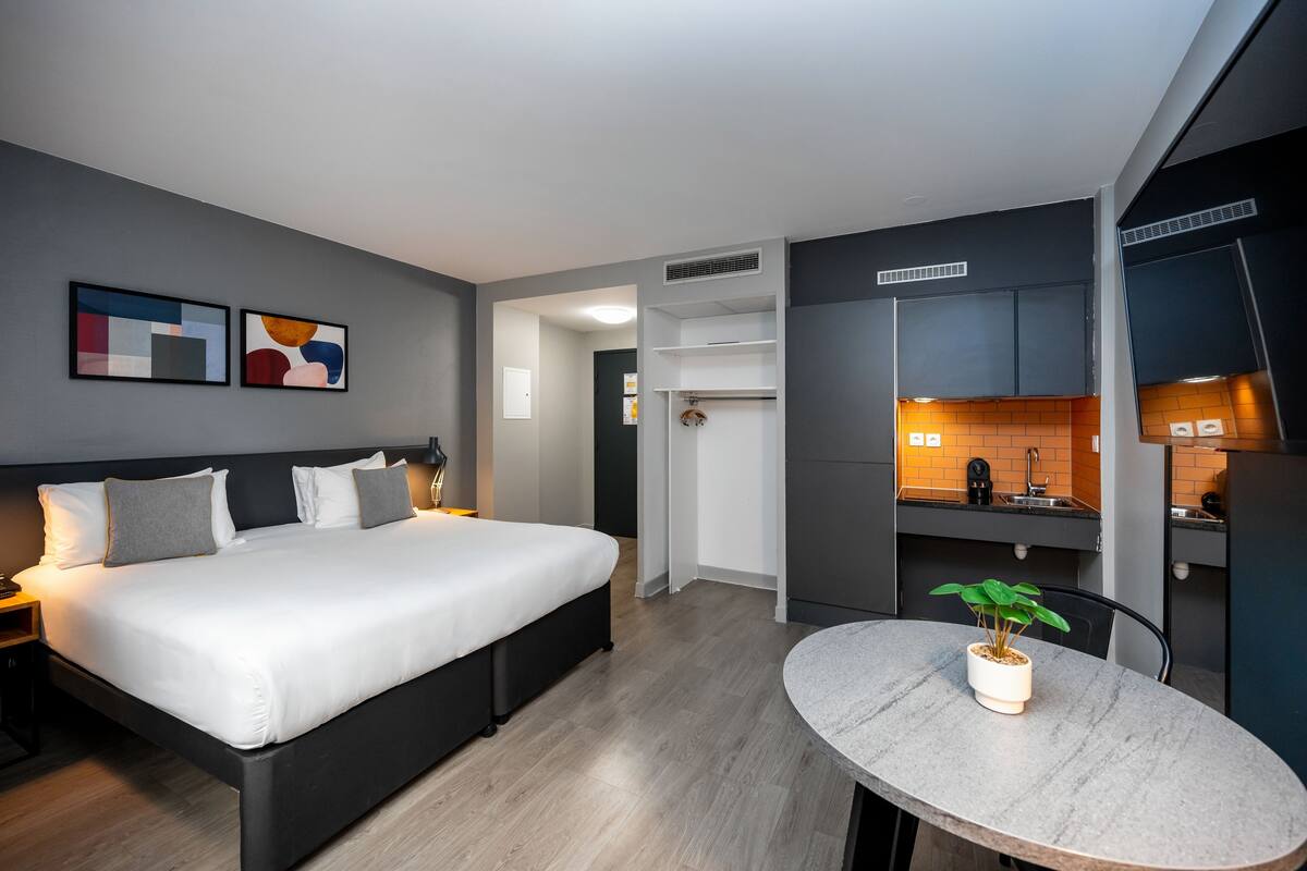 Une chambre d’hôtel moderne avec un grand lit, une petite table à manger, une plante en pot et une kitchenette.