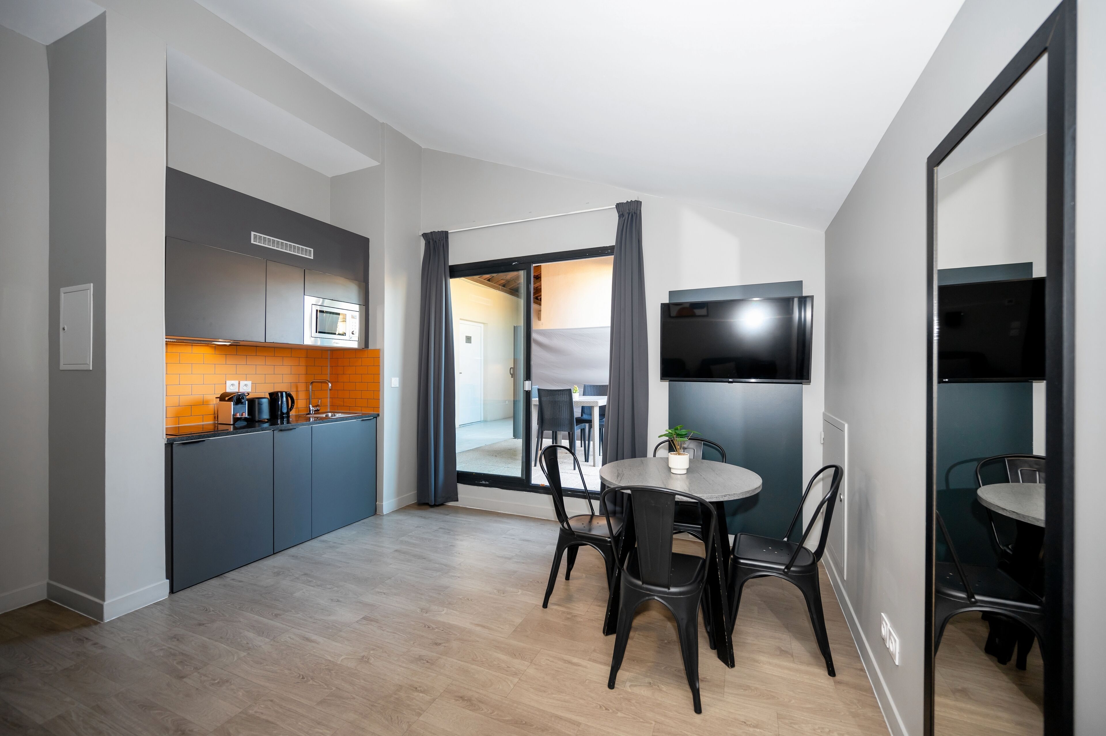 Cocina moderna con armarios grises, microondas y cafetera. Hay una zona de comedor con una mesa redonda y sillas, y un televisor montado en la pared.