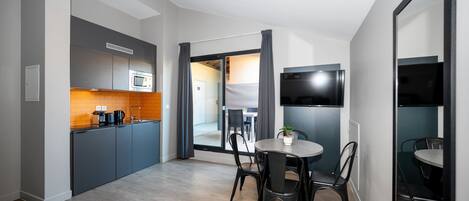 Cocina moderna con armarios grises, microondas y cafetera. Hay una zona de comedor con una mesa redonda y sillas, y un televisor montado en la pared.
