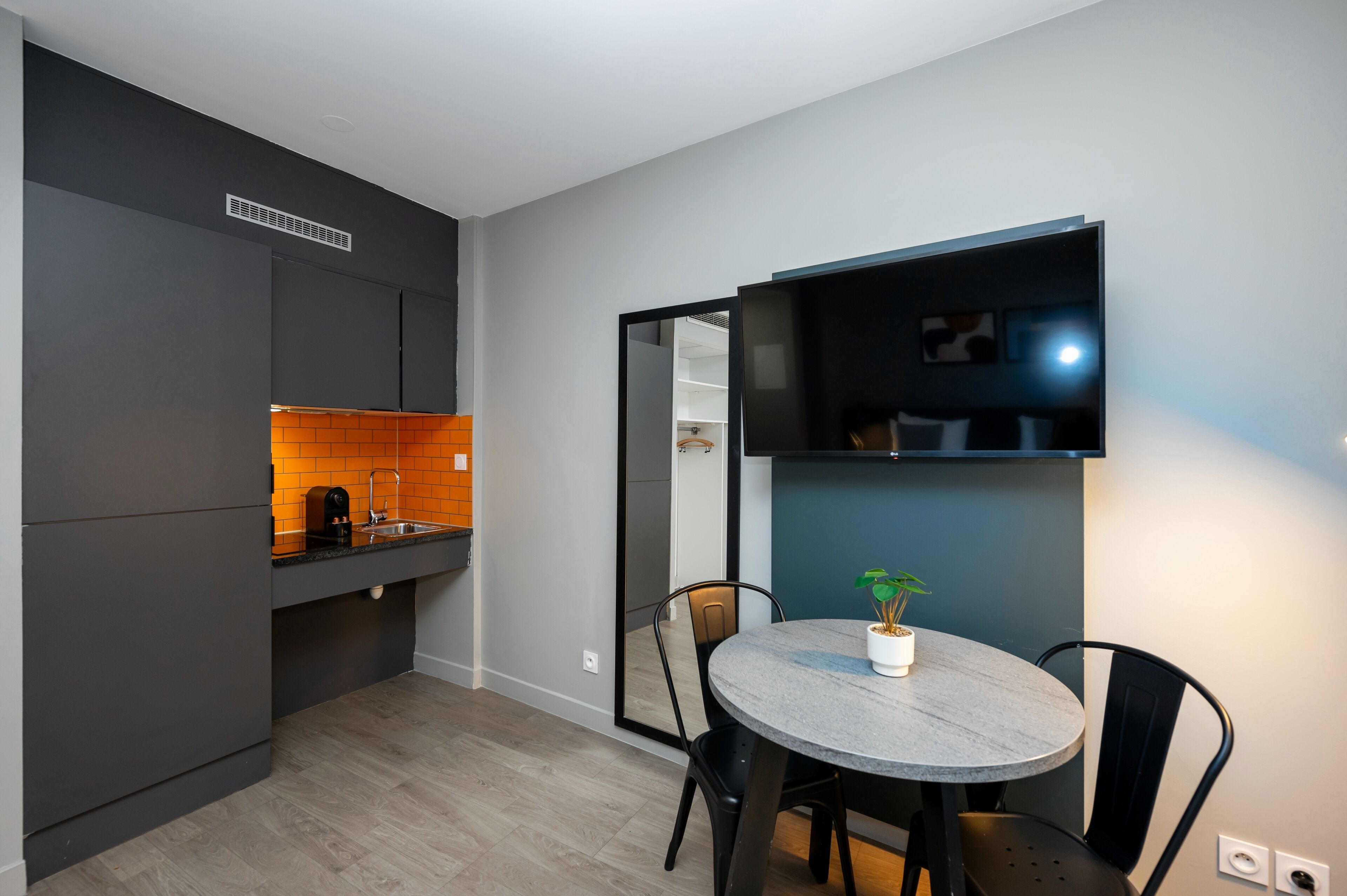 A modern kitchen with dark cabinetry, a built-in oven, and a sink. There is a dining area with a round table and chairs, and a potted plant on the table.