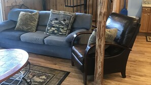 A living room with a blue sofa, a black leather chair, a wooden table, and a staircase. - Longmuir's Hidden Cove Cottage Resort (Addington Highlands)