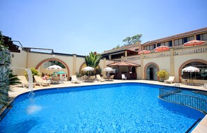 Une piscine entourée de chaises longues et de parasols, le tout bordé d’un bâtiment doté d’arcades et d’un toit en tuiles.