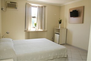 Une chambre à coucher avec un lit, une fenêtre avec des rideaux, un petit bureau avec une plante, un téléviseur fixé au mur et un radiateur.