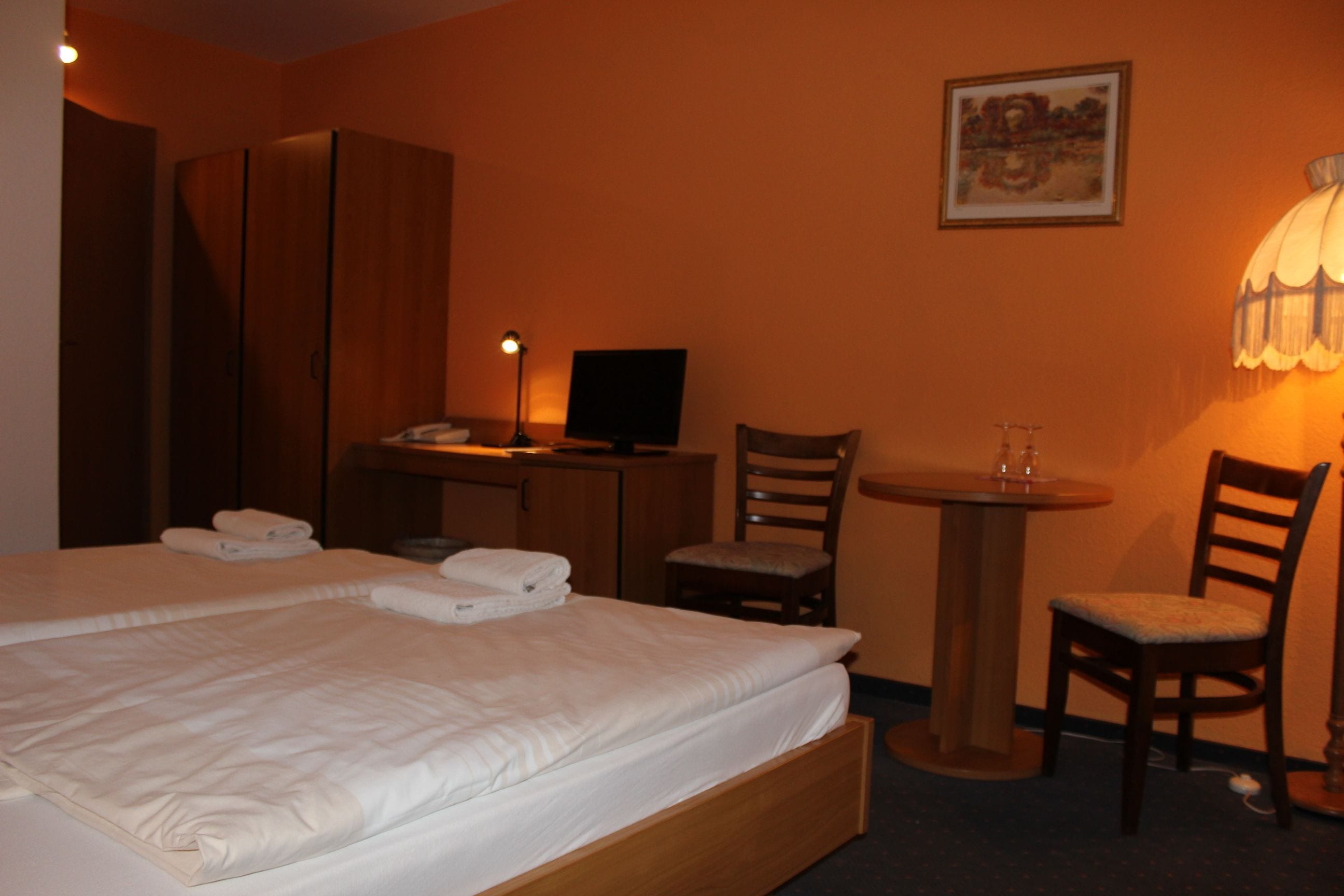 A hotel room with a bed, a desk with a computer, a chair, a small table with a lamp, and a painting on the wall.