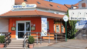 Un hotel con fachada roja, un cartel que indica 'Hotel Riedel' y un letrero de 'Biergarten' que señala un área de comedor al aire libre.