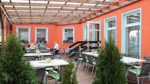 Un café al aire libre con mesas y sillas, cubierto por una pérgola de madera, y paredes de color naranja con ventanas.