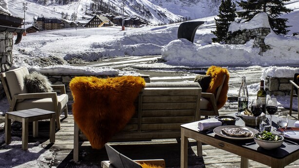 Un patio in legno con posti a sedere all'aperto, un tavolo apparecchiato per mangiare e uno sfondo montuoso con una seggiovia.