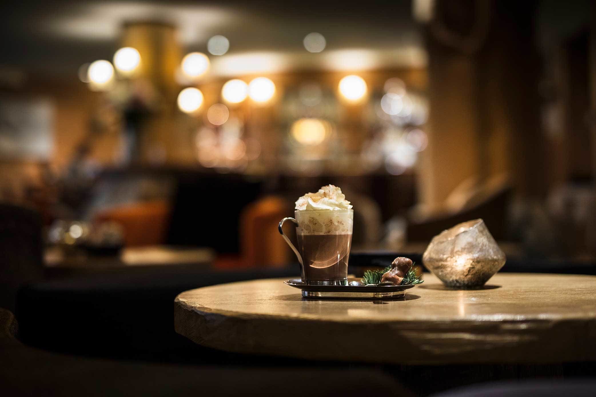 A cup of coffee with whipped cream on a wooden table in a dimly lit room with blurred background lights.