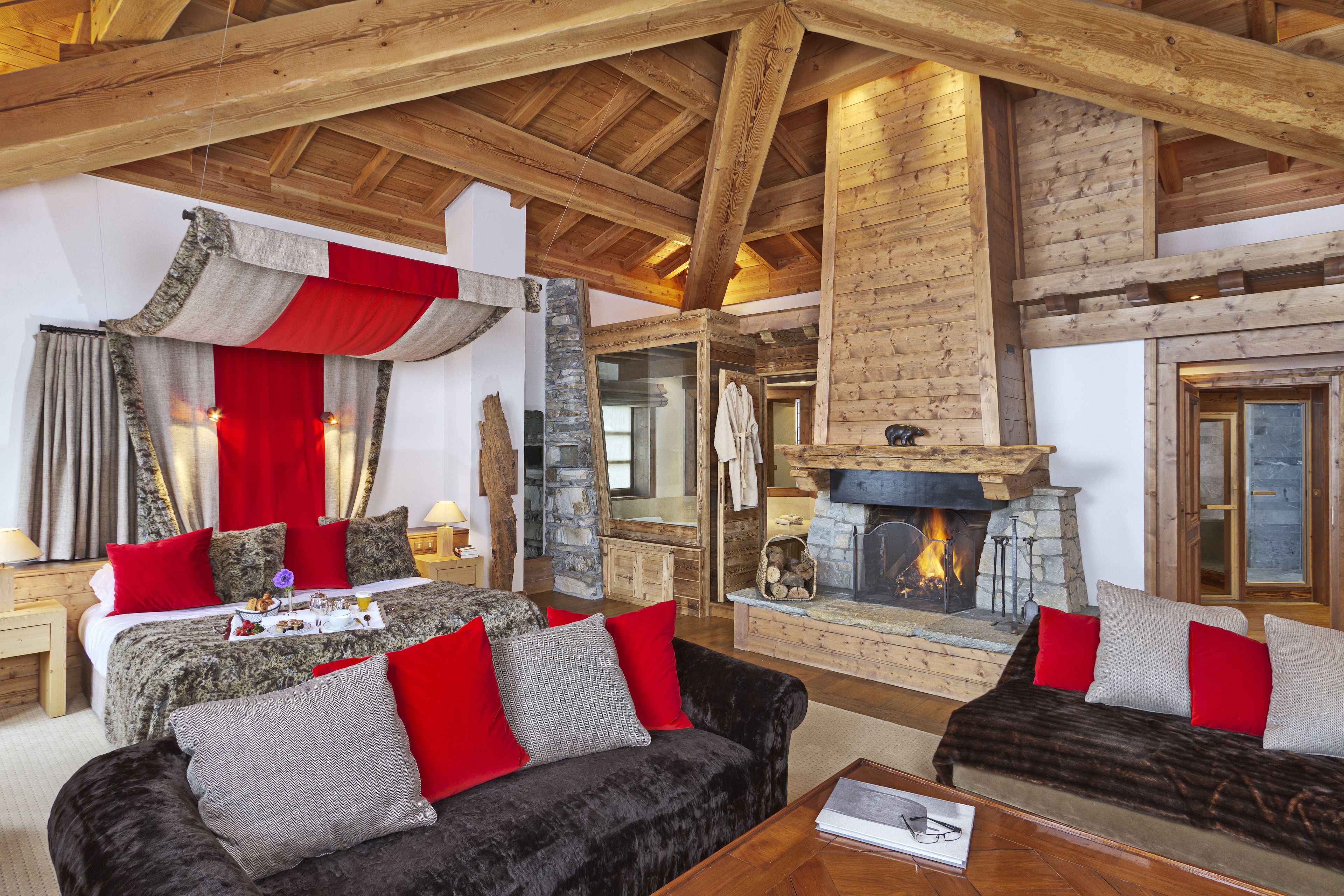 A spacious living area with a fireplace, a large bed with red and grey bedding, and a cozy seating area with red and grey cushions.