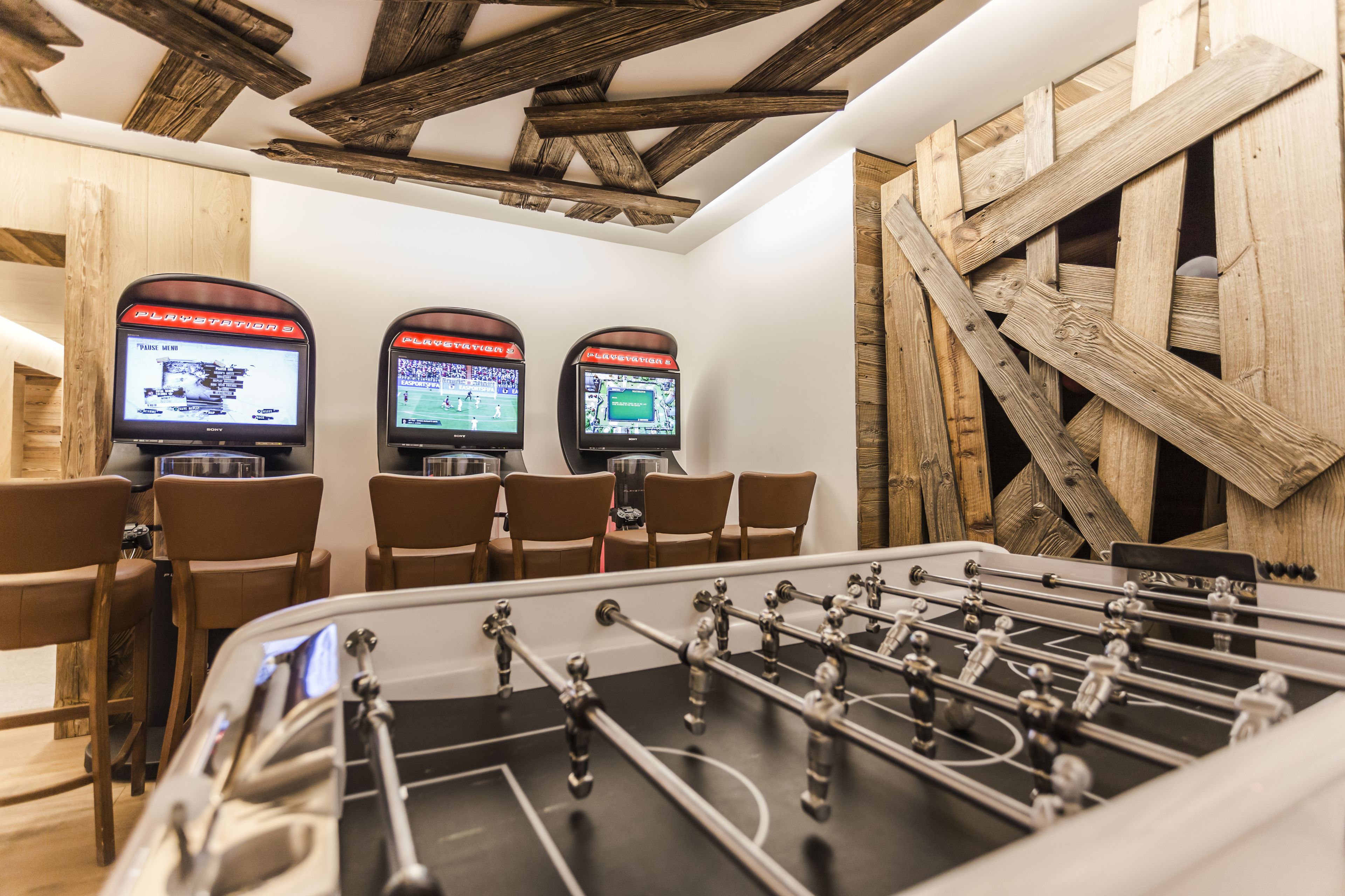 A room with a foosball table, two televisions mounted on the wall, and wooden chairs.