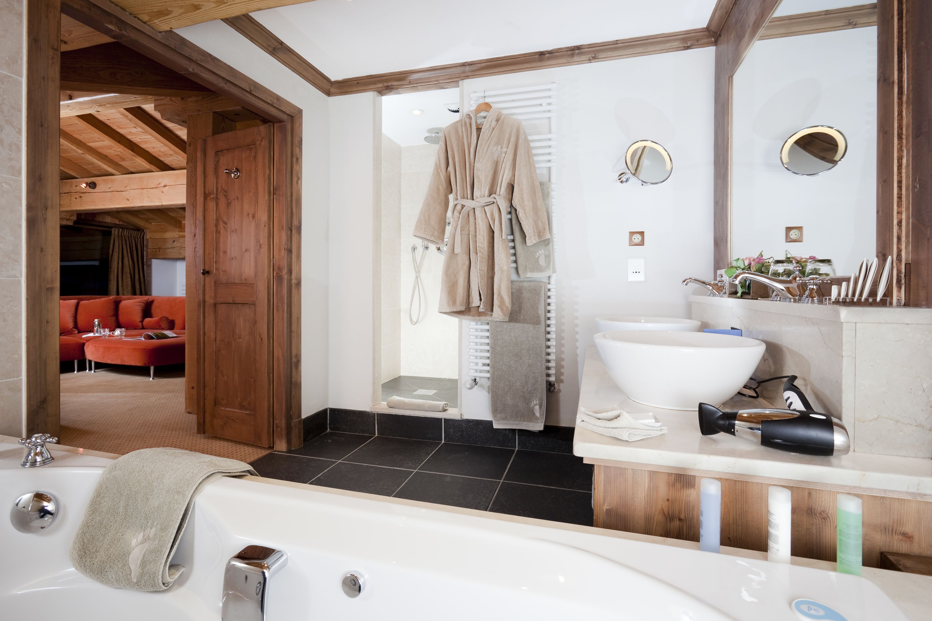 A modern bathroom with a bathtub, a large sink, and a shower. There is a wooden door leading to a living area with a red sofa.