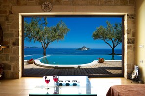 A room with a large window overlooking a pool with trees and a mountain view. - Kymaros Villas (Zakynthos)
