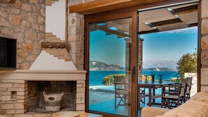 Una chimenea de piedra con una mesa de madera y una cesta de mimbre, una puerta de cristal que da a una zona de estar exterior con vistas al mar y a las montañas.