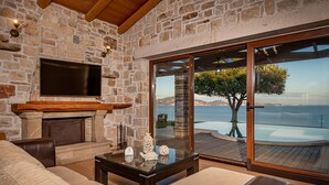 Un salón con paredes de piedra, una chimenea, un televisor de pantalla plana y un amplio ventanal que da a una terraza con vistas al mar.