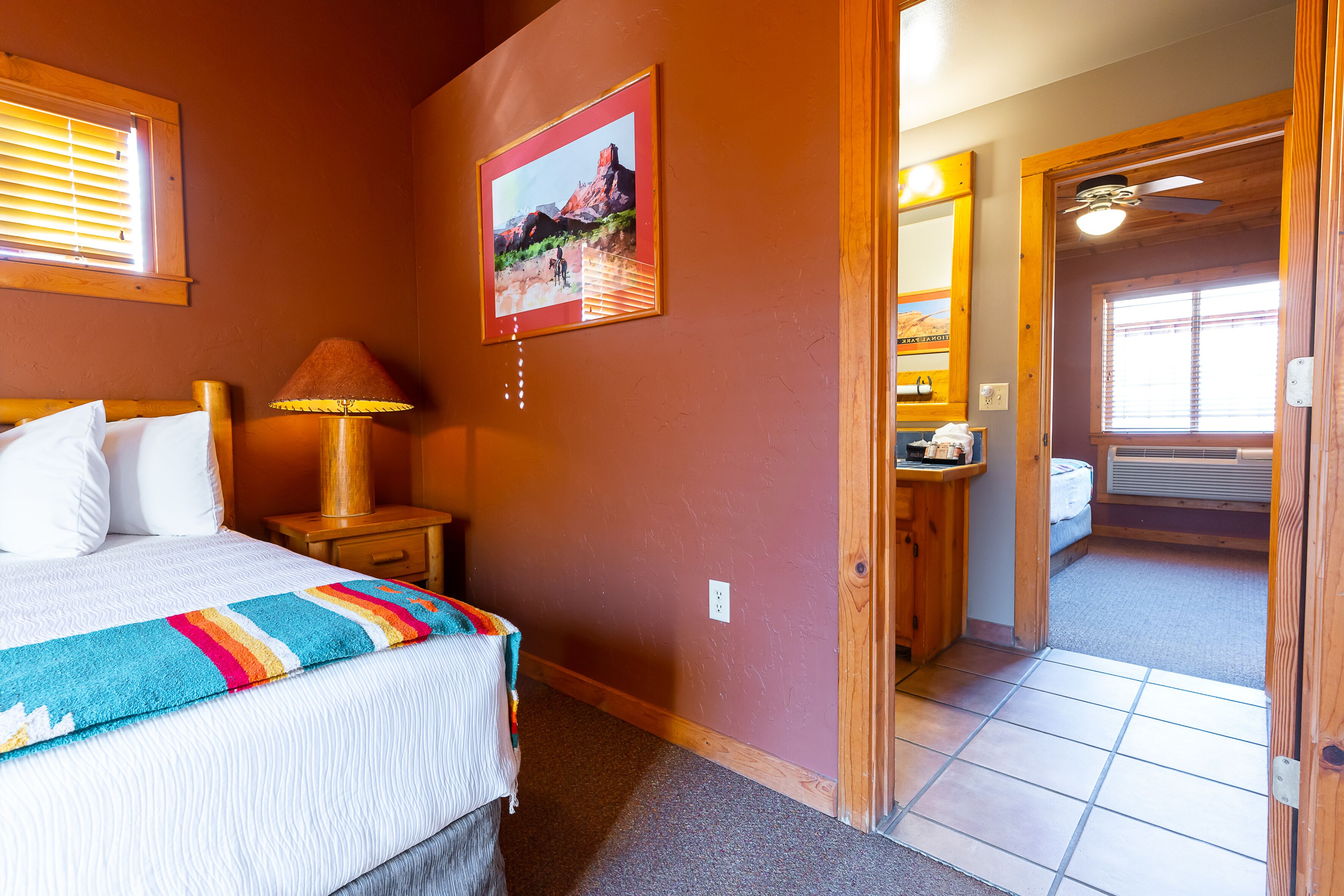 A bedroom with a bed, a nightstand, a lamp, a painting, and a door leading to another room.