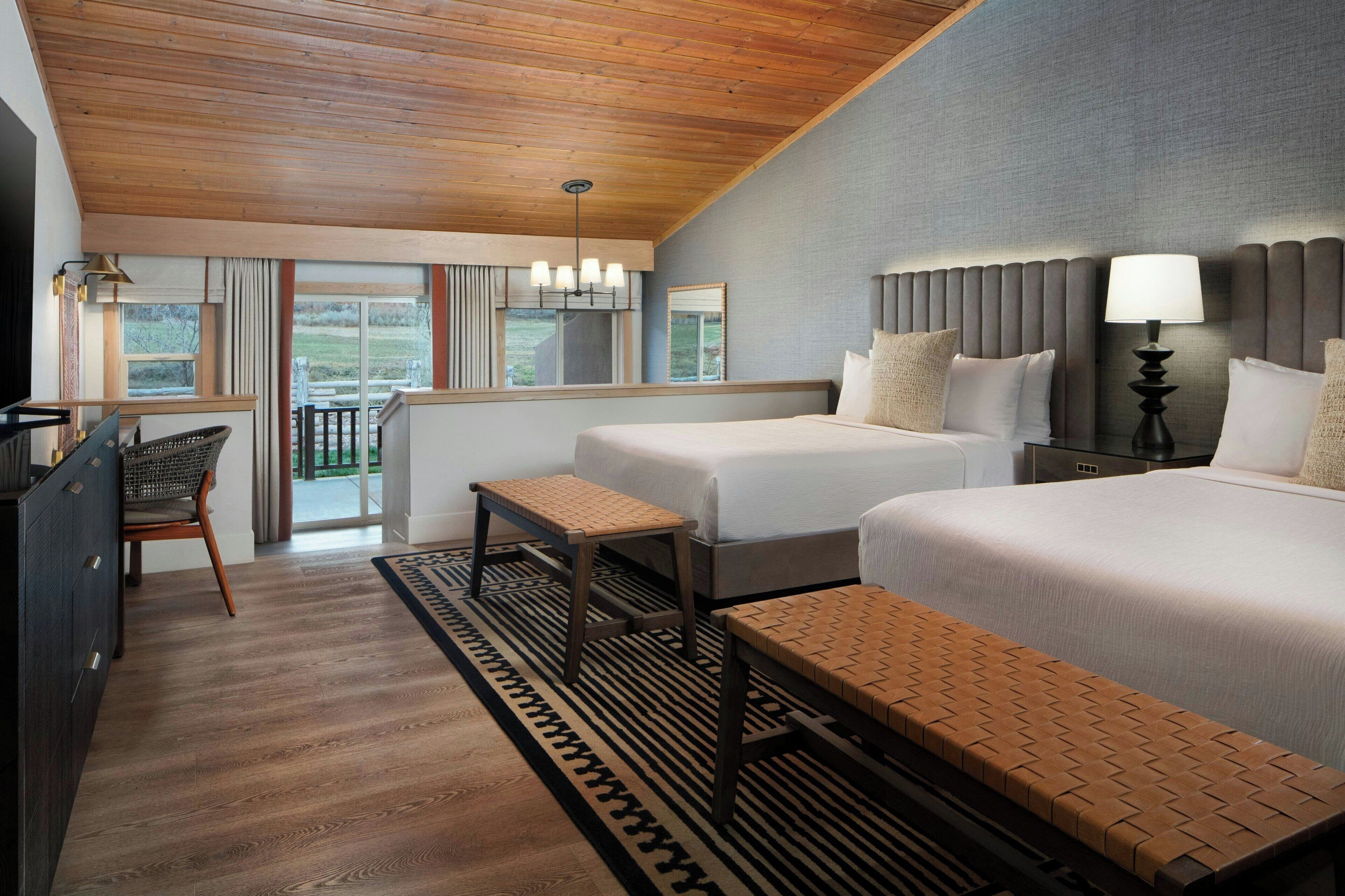 A hotel room with two beds, a desk, a chair, and a view of the outdoors.