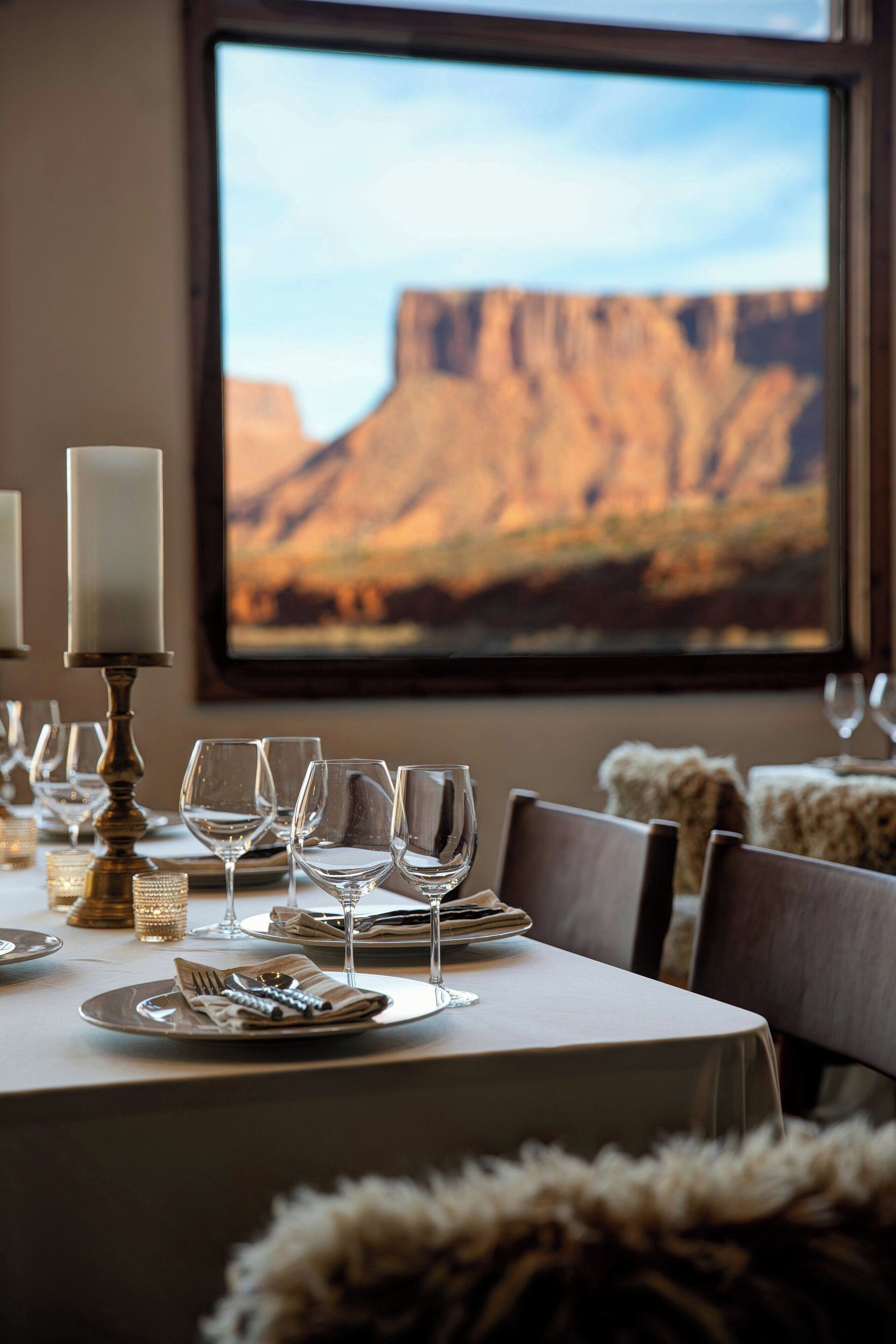 A dining table set with plates, glasses, and cutlery, with a candle holder and a view of a mountainous landscape through a window.