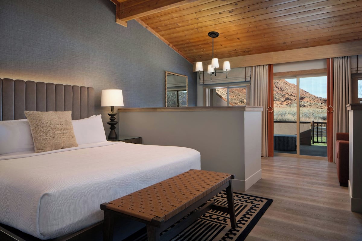 A bedroom with a large bed, a bedside table with a lamp, a bench, a mirror, and a view of a mountainous landscape through a sliding door.