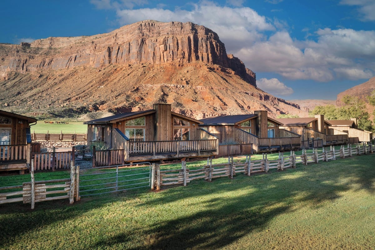 A rustic lodge with wooden cabins, a fenced yard, and a mountain backdrop.