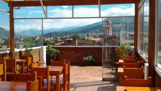 Un área de estar al aire libre con techo, mesas y sillas de madera, vistas a un paisaje urbano y montañas al fondo, y un recinto de vidrio que separa la zona de estar de las vistas.