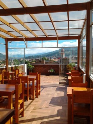 A covered outdoor seating area with wooden tables and chairs, a view of a cityscape and mountains in the background, and a glass enclosure separating the seating from the view. - El Jacal Classic (Huaraz)