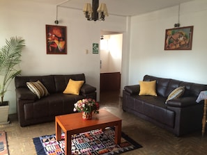 A room with two dark brown sofas, each with a yellow pillow, a wooden coffee table with a colorful vase, and a patterned rug on a wooden floor. - El Jacal Classic (Huaraz)