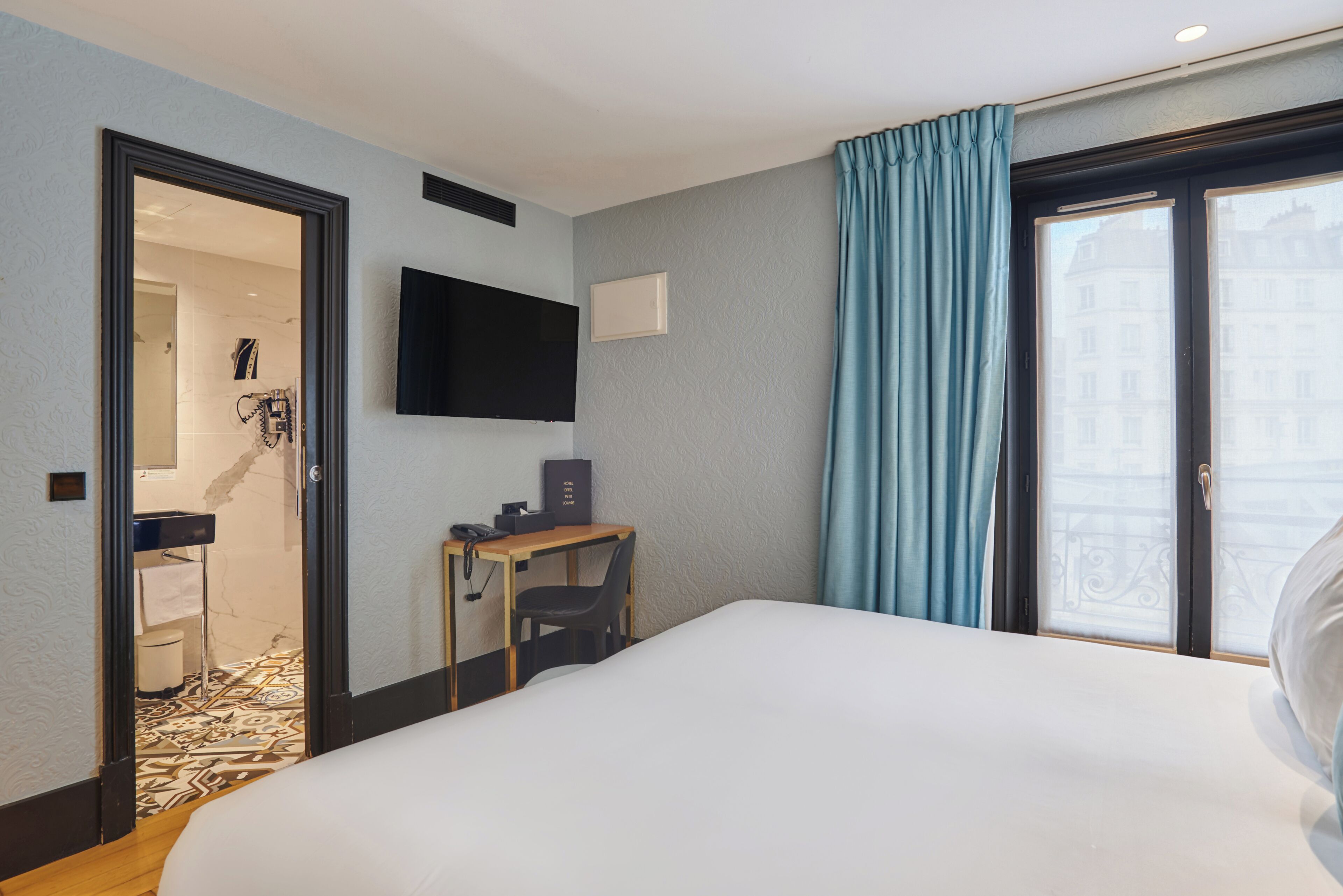 A hotel room with a bed, a desk with a chair, a flat-screen TV, and a bathroom visible through an open door.