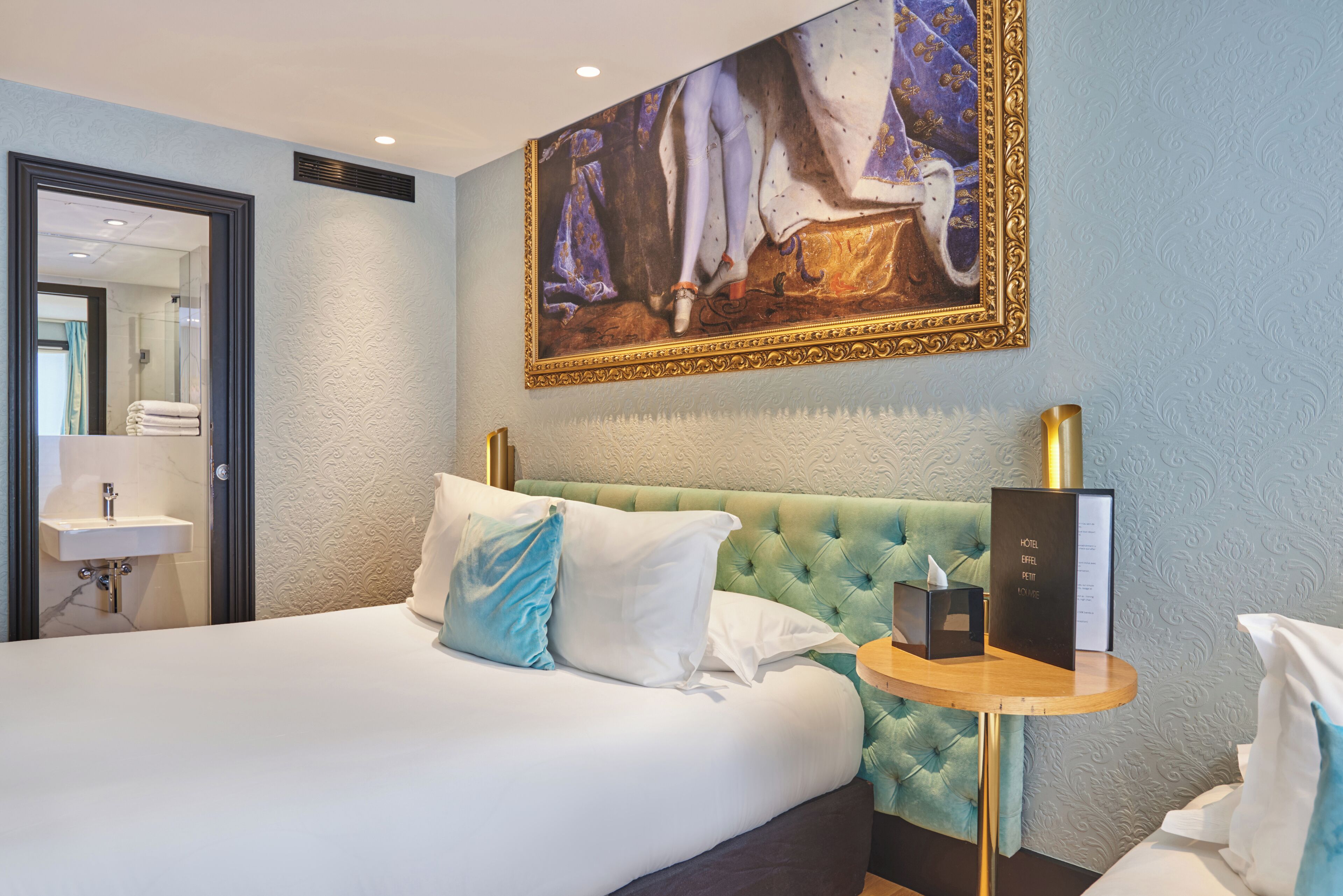 A hotel room with a bed, a round wooden side table, a wall-mounted painting, a sink, and a mirror.