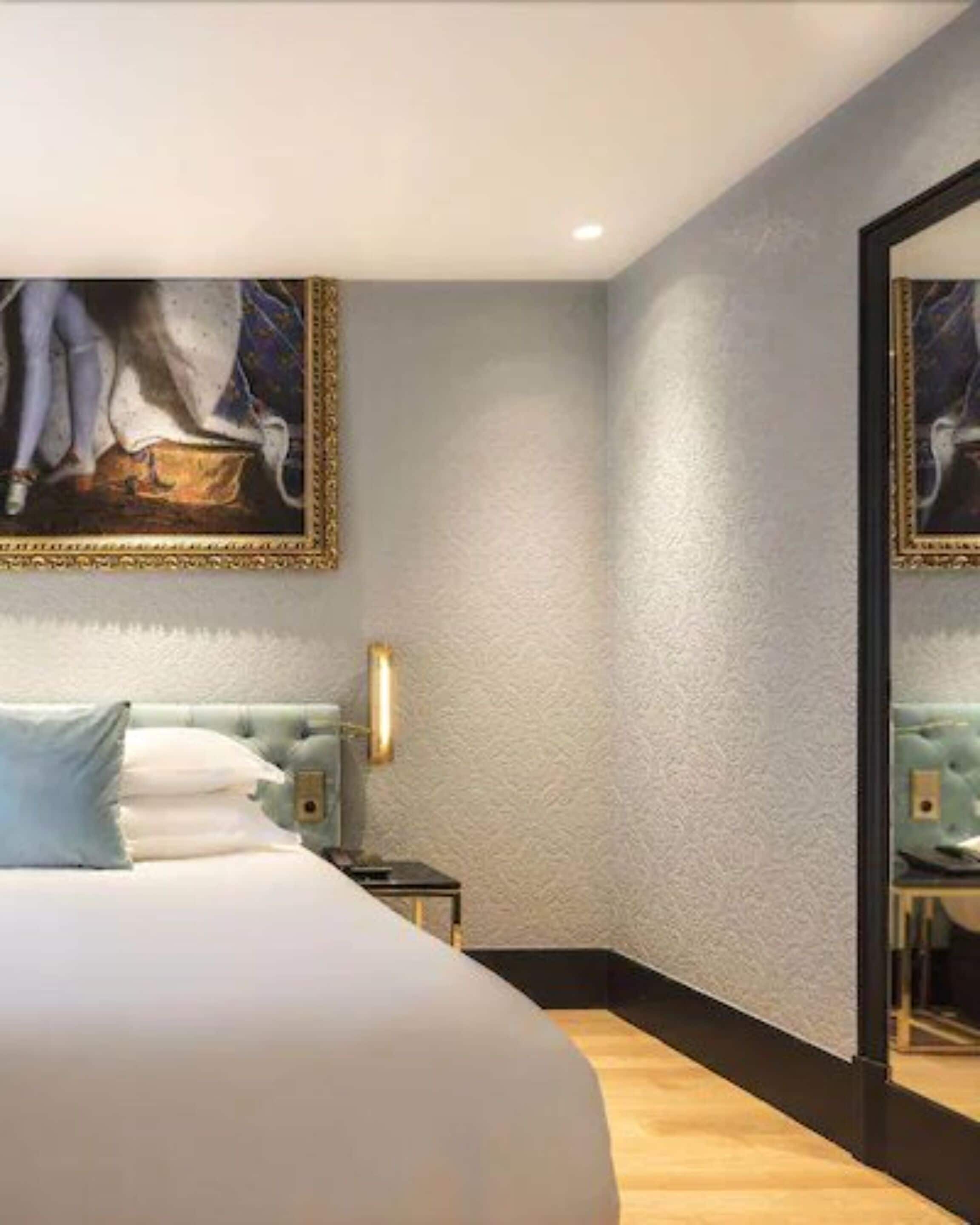 A modern hotel room with a large bed, a nightstand, a wall-mounted lamp, and a large mirror.