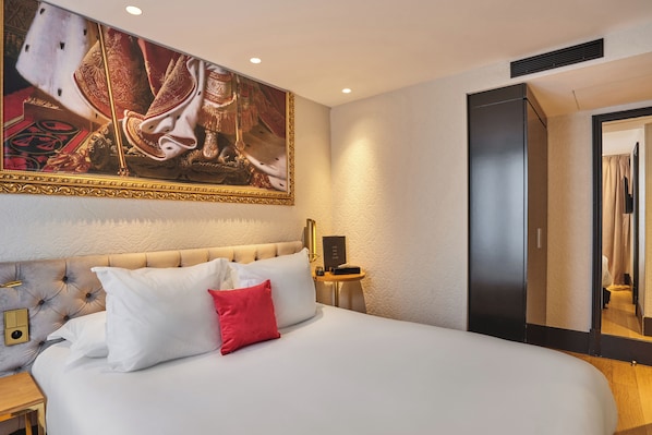 A hotel room with a large bed, a headboard with tufting, a red accent pillow, a wall-mounted painting, a bedside table with a lamp, and a door with a mirror. - Hôtel Eiffel Petit Louvre (Paris)