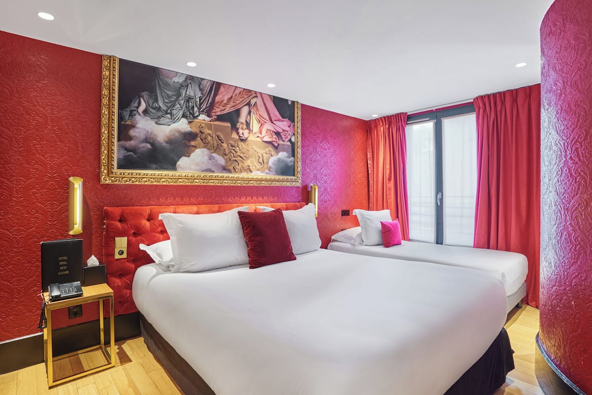 Una habitación de hotel con dos camas, paredes rojas, un cuadro grande y una ventana con cortinas rojas.